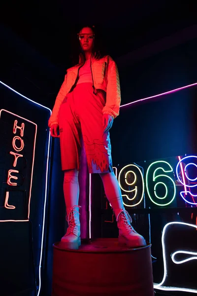 Low angle view of asian woman in sunglasses looking at camera while posing near red neon hotel sign — Photo de stock