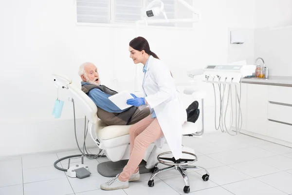 Sorprendido paciente mayor acostado en la silla dental y tomando la consulta del médico con el comprimido en las manos - foto de stock