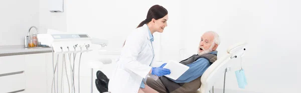 Dentista sorrindo mostrando tablet para paciente sênior espantado na clínica odontológica, banner — Fotografia de Stock