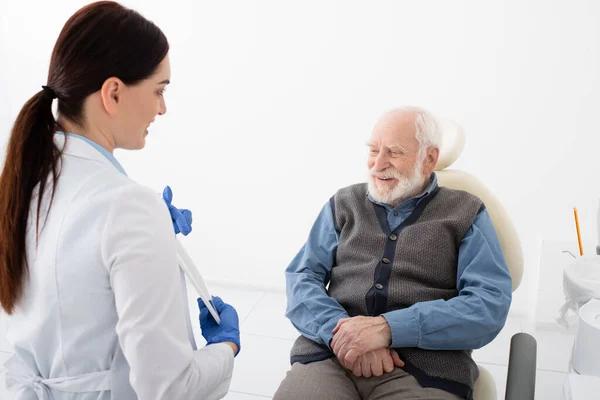 Patient âgé dans une chaise dentaire en consultation avec un médecin souriant avec un comprimé dans les mains — Photo de stock