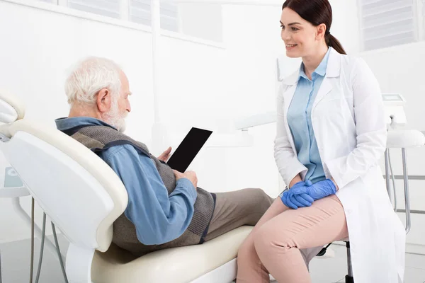 Senior patient lying in dental chair and holding tablet with blank screen in hands near doctor in clinic — Stock Photo