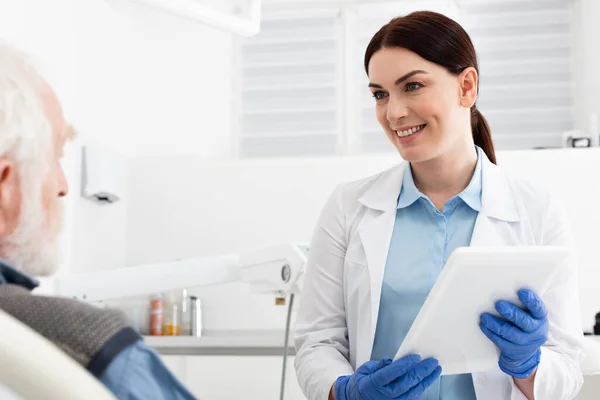 Dentista alegre con la tableta en las manos mirando al paciente mayor sentado en la silla dental - foto de stock