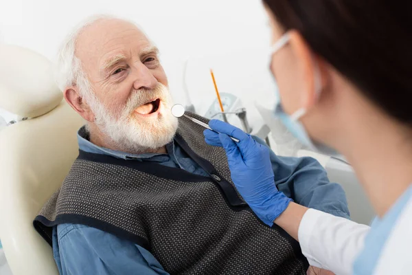 Пожилой человек с осмотром зубов стоматологом в латексных перчатках с зеркалом в стоматологическом кресле — стоковое фото