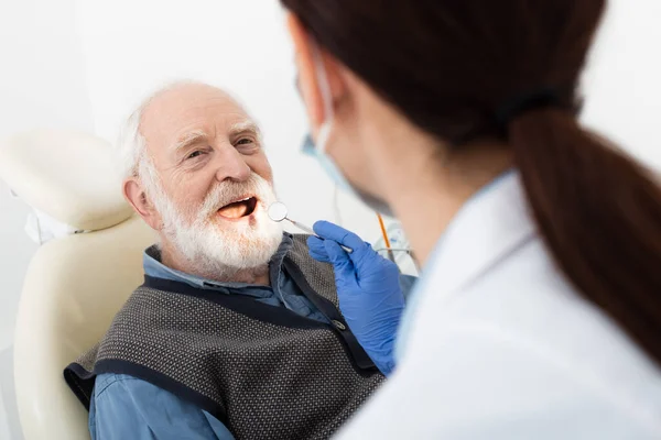 Hombre mayor que tiene examen de dientes por dentista en guantes de látex con espejo en la mano en silla dental - foto de stock