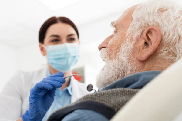 Dentista en máscara médica examinar los dientes de hombre mayor con sonda y espejo en la silla dental - foto de stock