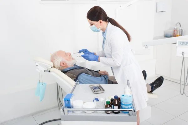 Dentista examinando dentes de homem idoso com sonda e espelho na clínica odontológica — Fotografia de Stock