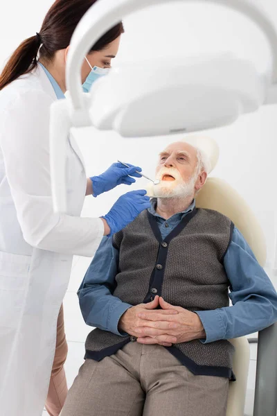 Стоматолог, осматривающий зубы пожилого человека с зондом и зеркалом в стоматологической клинике — стоковое фото