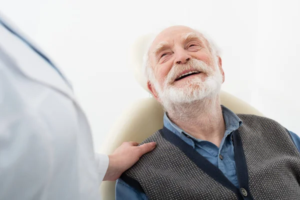 Усміхнений старший пацієнт сидить у стоматологічному кріслі з рукою стоматолога на плечі — стокове фото