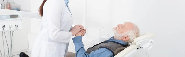 Patient âgé allongé dans la chaise du dentiste et tenant la main avec le dentiste, bannière — Photo de stock