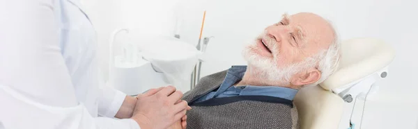Patient âgé reconnaissant dans une chaise dentaire tenant la main avec un dentiste, bannière — Photo de stock