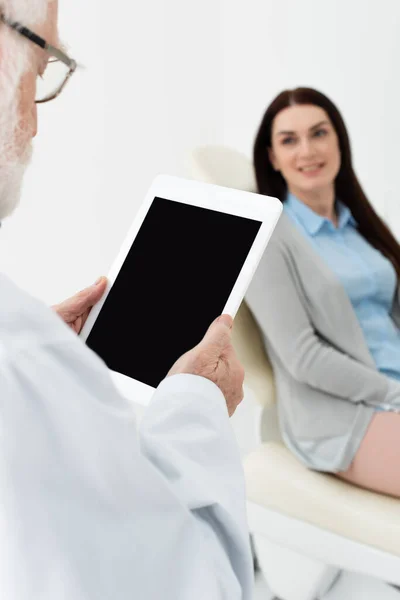 Dentista senior de pie con la tableta de pantalla en blanco delante de la mujer borrosa en la silla dental en la clínica - foto de stock