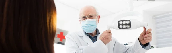 Dentista senior en máscara médica tocando lámpara médica delante del paciente, bandera - foto de stock