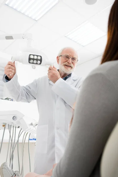 Alegre dentista senior tocando lámpara médica delante de la mujer en silla dental - foto de stock