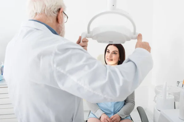 Dentista senior que regula la lámpara médica delante del paciente en silla dental - foto de stock