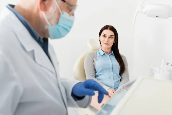 Позитивная женщина сидит в стоматологическом кресле и смотрит на стоматолога, стоящего рядом с оборудованием в клинике — стоковое фото