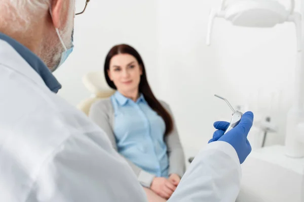 Dentiste chevronné avec équipement médical en main devant le patient en fauteuil dentaire — Photo de stock