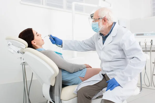 Dentiste senior en masque médical faisant le traitement des dents du patient dans la chaise dentaire — Photo de stock