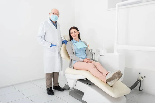 Dentiste senior en masque médical et gants en latex debout près de jolie femme assise dans une chaise dentaire à la clinique — Photo de stock