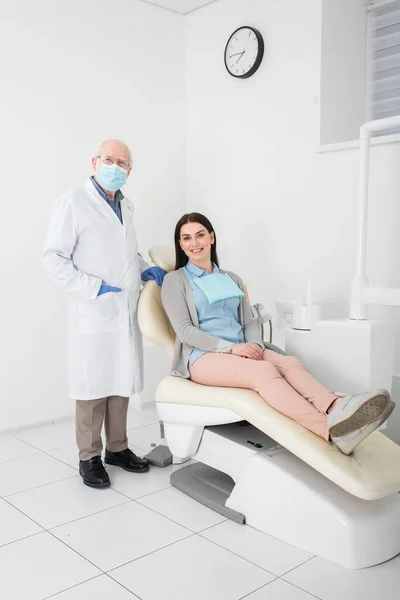 Dentista senior en bata blanca, máscara médica y guantes de látex de pie cerca del paciente en silla dental - foto de stock