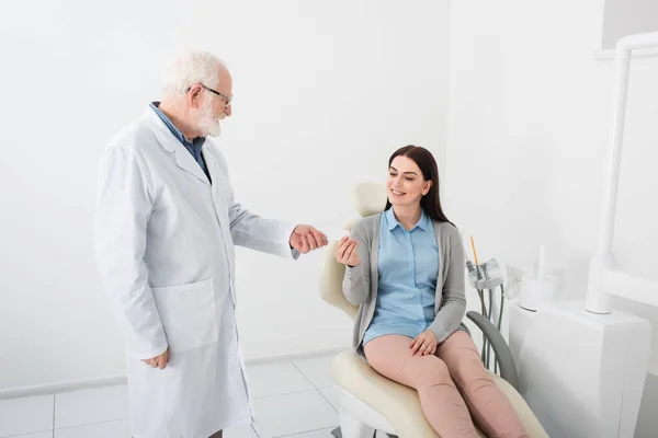 Senior dentist giving empty card to smiling woman in dental chair in clinic — Stock Photo