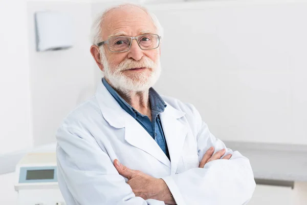 Ritratto di dentista anziano in mantello bianco e occhiali in piedi con le braccia incrociate in clinica dentale — Foto stock