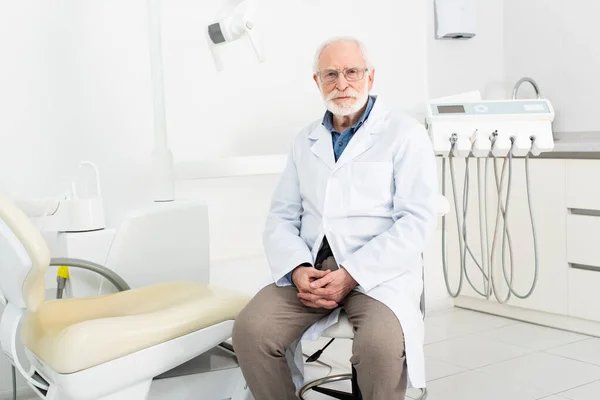 Dentista senior con abrigo blanco sentado con las manos cerradas en la clínica dental - foto de stock