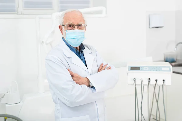 Dentiste senior en manteau blanc et masque médical debout avec bras croisés en clinique dentaire — Photo de stock