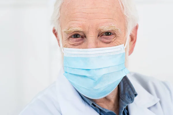 Portrait of senior doctor in white coat and medical mask on grey background — Stock Photo