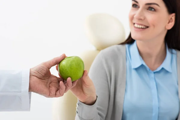 Vista de cerca de la manzana verde en las manos de la mujer adulta sonriente y el médico en la clínica dental - foto de stock