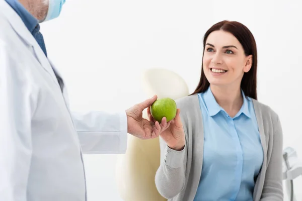 Mujer adulta sonriente en silla dental y médico sosteniendo manzana verde en la clínica - foto de stock
