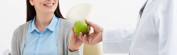 Cropped view of smiling adult woman in dental chair and doctor holding green apple in clinic, banner — Stock Photo