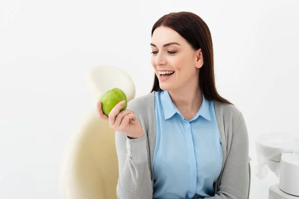 Mujer adulta sonriente en silla dental mirando manzana verde en la mano en la clínica - foto de stock