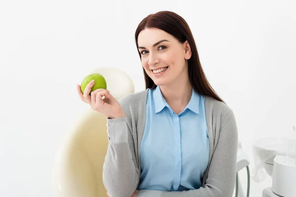 Smiling adult woman in dental chair holding green apple and looking at camera in hand in clinic — Stock Photo