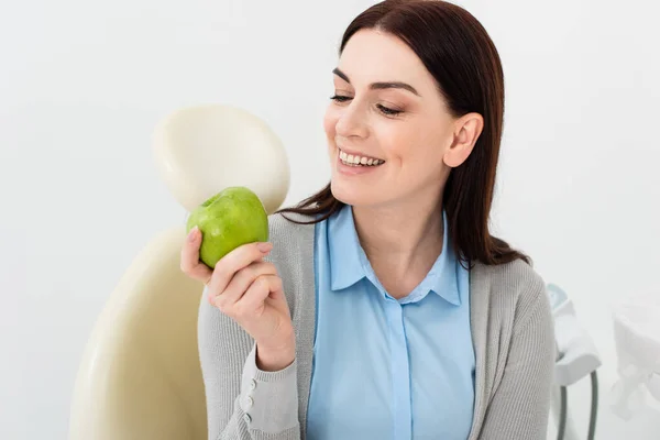 Smiling adult woman in dental chair holding green apple in hand in clinic — Stock Photo