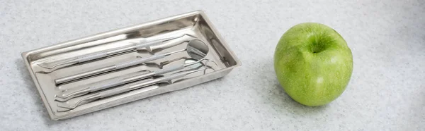 Vue grand angle des outils dentaires en métal dans le plateau et pomme verte sur la table médicale, bannière — Photo de stock