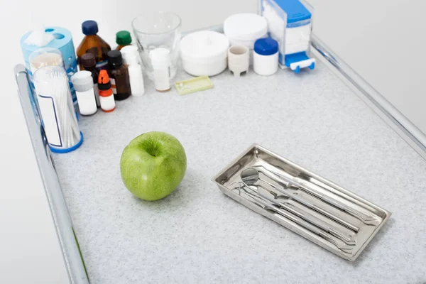 Высокий угол обзора лекарств и стоматологических инструментов в металлическом лотке и зеленом яблоке на медицинском столе — стоковое фото
