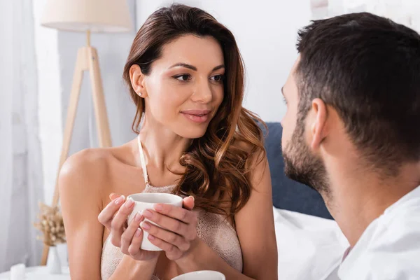 Pretty woman in bra holding cup near blurred boyfriend in bedroom — Stock Photo