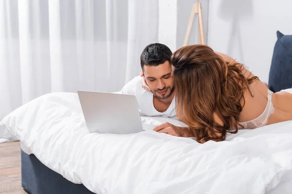 Mujer en sujetador besar novio cerca de portátil en la cama - foto de stock