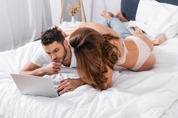 Молодая женщина в нижнем белье трогает парня с помощью ноутбука на кровати — стоковое фото