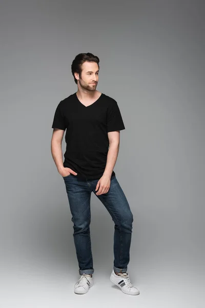Intera lunghezza dell'uomo barbuto in jeans e t-shirt nera in posa con mano in tasca su grigio — Foto stock