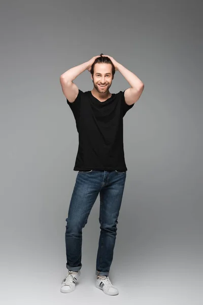 Comprimento total do homem barbudo em jeans e camiseta preta posando e sorrindo em cinza — Fotografia de Stock