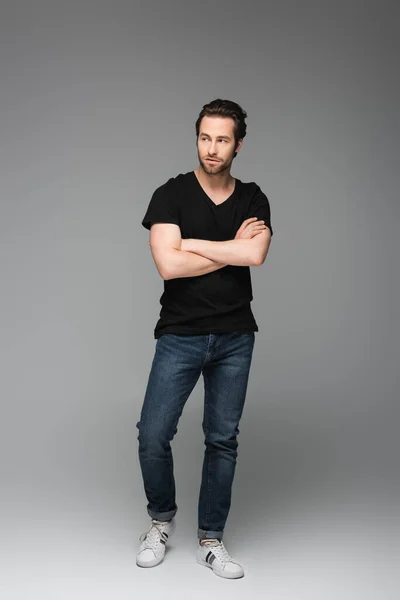 Comprimento total do homem barbudo em jeans e camiseta preta posando com braços cruzados em cinza — Fotografia de Stock