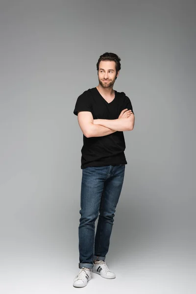Intera lunghezza di bell'uomo in jeans e t-shirt nera in posa con le braccia incrociate sul grigio — Foto stock