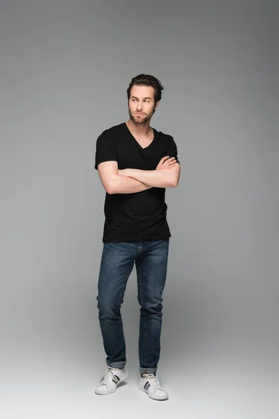 Hombre barbudo serio en jeans y camiseta negra posando con los brazos cruzados sobre gris - foto de stock