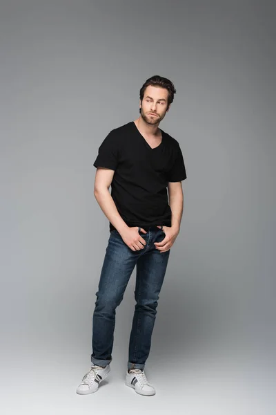 Tutta la lunghezza dell'uomo barbuto in jeans e t-shirt nera in posa mentre distoglie lo sguardo sul grigio — Foto stock