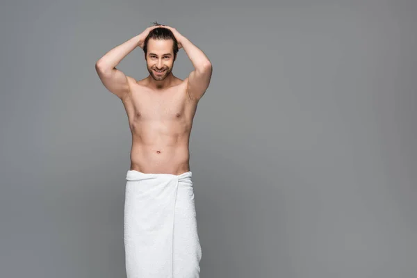 Hombre musculoso feliz envuelto en toalla ajustando el cabello aislado en gris - foto de stock
