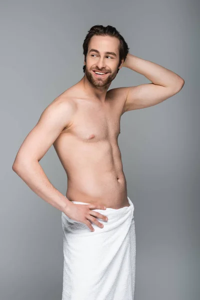 Happy and muscular man wrapped in white towel standing with hand on hip isolated on grey — Stock Photo