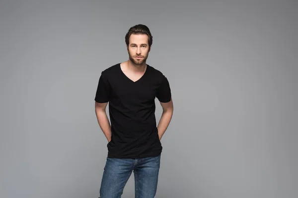 Hombre barbudo en camiseta negra y jeans posando con las manos detrás de la espalda aisladas en gris - foto de stock