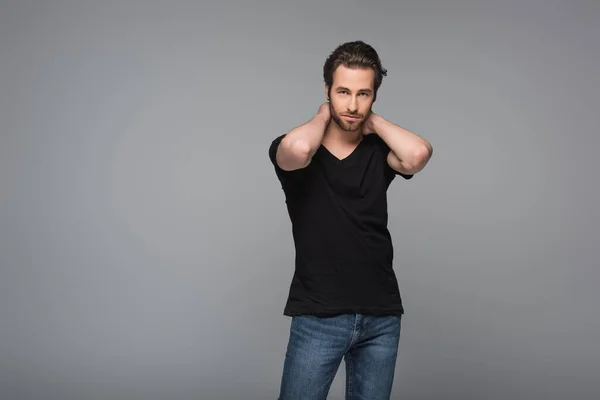 Bearded man in black t-shirt and jeans posing with hands near neck isolated on grey — Stock Photo