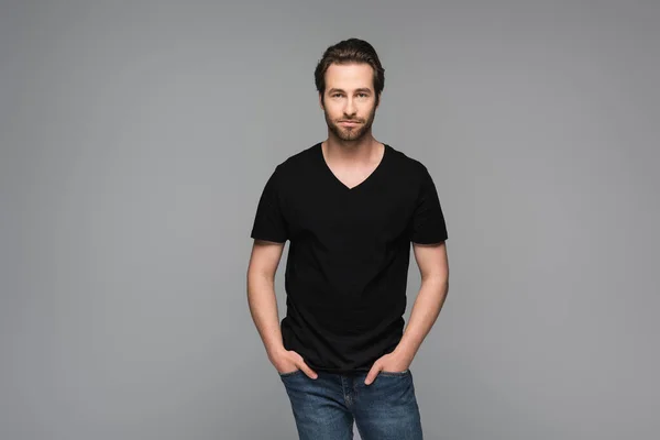 Hombre barbudo en camiseta negra y jeans posando con las manos en bolsillos aislados en gris - foto de stock